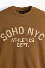 Buzo marrón de algodón con cuello redondo, hombros caídos y diseño estampado en el frente con la inscripción 'SOHO NYC ATHLETICS DEPT.' en color beige. Presenta puños y bajo de canalé elástico.