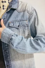 Camisa de denim azul con lavado gastado, bolsillos con solapa en el pecho, cuello en punta y cierre frontal de broches metálicos.