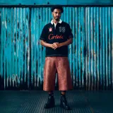 Remera polo de manga corta de la marca Corteiz, color negro, con cuello blanco y cierre de botones oculto. Presenta detalles bordados y estampados, incluyendo el texto 'Corteiz' en rojo en el pecho, el número '18' y diversos parches en las mangas.