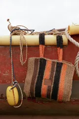 Cartera tipo tote de diseño tejido a rayas, combinando tonos beige, marrón, negro y naranja. Cuenta con doble asa superior y detalle de etiqueta frontal.