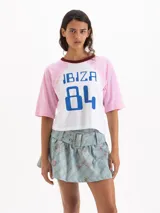 Remera estilo raglán de manga corta, con cuerpo blanco, mangas rosadas y cuello redondo bordó. Cuenta con un estampado frontal en azul con las palabras 'IBIZA 84' y detalles de encaje en las costuras de las sisas.