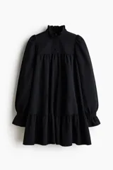 Vestido corto de corte holgado con cuello alto de volados y cierre frontal de botón. Presenta canesú fruncido, mangas largas abullonadas con puños acampanados y falda con costura fruncida en el bajo.