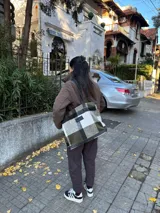 Bolso tipo tote de gran capacidad con diseño de cuadros en tonos verde oliva, blanco y negro. Presenta detalles en cuero negro en la base y en las correas de hombro, además de un parche de cuero con logo en el frente. Incluye cierre superior y bolsillo interior.