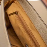 Bandolera de material símil cuero color taupe, con solapa frontal redondeada y cierre superior. Posee correa larga regulable y desmontable, y herrajes metálicos dorados. Lleva un logo metálico de la marca XL aplicado al frente.