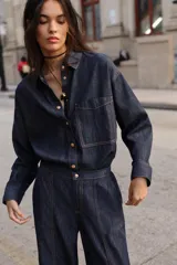 Camisa amplia de denim azul oscuro con cuello solapa, manga larga, bolsillo de plastrón en el pecho y cierre frontal con botones a presión dorados.