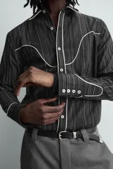 Camisa de corte relajado en color negro con rayas verticales finas. Presenta un diseño estilo western con vivos blancos en contraste en el canesú, cuello, tapeta frontal y puños. Cuenta con cuello solapa, cierre frontal de botones y mangas largas.