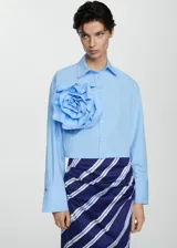 Camisa crop de color celeste con cuello camisero, manga larga con puños abotonados y cierre frontal de botones. Destaca por una maxi flor decorativa aplicada en el pecho.