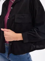 Campera de pana negra con cierre frontal de botones metálicos, cuello clásico y dos bolsillos frontales con solapa. Presenta mangas largas con puños abotonados y un diseño liviano sin forro.