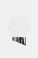 Camiseta oversize de cuello redondo y manga corta, confeccionada en algodón blanco. Presenta un bajo asimétrico con una aplicación de lentejuelas negras brillantes en uno de los laterales.