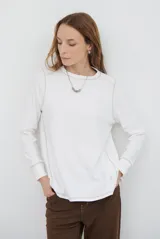 Remera de manga larga en color beige, confeccionada en tejido de punto con costuras a la vista en contraste. Presenta un corte holgado, cuello redondo y un pequeño bordado distintivo en la parte inferior.