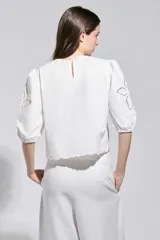 Top Allegra de color blanco, con mangas abullonadas a la altura del codo y puños elásticos. Presenta detalles de calados y bordados florales en los hombros, mangas y ruedo.