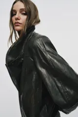 Chaqueta de corte amplio estilo biker en tejido efecto piel de color verde oscuro, con cuello alto envolvente, cierre frontal asimétrico de cremallera metálica, bolsillos ocultos en costura y forro interior.
