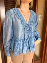 Blusa de tela satinada color azul, con escote en V, volados decorativos en el frente y mangas largas abullonadas con puños ajustados. Presenta un lazo frontal para atar y un diseño con volado en el ruedo.