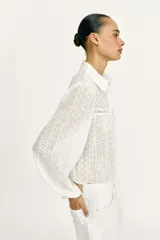 Blusa corta de color blanco con diseño de bordados perforados, cuello solapa, cierre frontal con botones y mangas largas abullonadas.