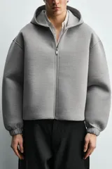 Sudadera gris de corte boxy fit con capucha y cierre frontal de cremallera. Confeccionada en tejido doble y compacto, cuenta con bolsillos laterales ocultos en la cadera y mangas largas con puños elásticos.
