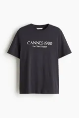 Remera de algodón de corte holgado, color gris oscuro, con cuello redondo y estampado frontal con la leyenda 'CANNES 1980 La Côte D'azur'.