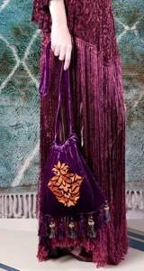 Cartera de mano tipo bolsa confeccionada en terciopelo de seda color violeta, con bordado floral en hilo dorado y detalle de borlas colgantes.