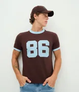 Remera marrón de manga corta con cuello redondo y ribetes celestes en cuello y puños. Presenta un gran estampado numérico "86" en color celeste en el pecho.