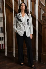 Blazer de corte sastrero con estampado de rayas verticales en tonos gris y blanco. Presenta una intervención artística en el pecho con apliques de cuero, encaje, tachas y piedras, además de un detalle de borla negra. Cuenta con cierre frontal de un botón y solapas clásicas.