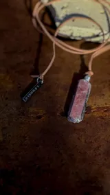 Collar largo con tiras de cuero sintético tostado y dije de piedra en bruto de tonalidad rojiza con detalles metálicos, acompañado de una placa metálica grabada.