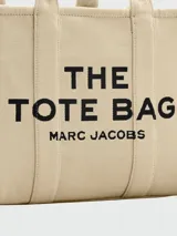 Bolso tipo tote de Marc Jacobs, modelo The Jacquard Large Tote Bag, confeccionado en tejido jacquard color tostado. Presenta el nombre de la marca y el modelo bordados en el frente en color blanco, doble asa superior y detalles de cintas laterales con inscripciones.