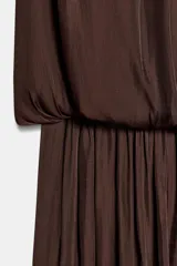 Vestido corto de tejido satinado color marrón, con cuello redondo, mangas largas con diseño fluido y cintura elástica. Presenta una abertura frontal y cierre posterior con botón.