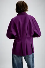 Camisa de manga larga en color violeta, con cuello solapa y cierre frontal de botones. Presenta un diseño de corte recto con detalle de pliegues en la espalda.