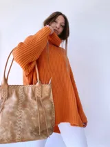 Cartera tipo tote de cuero color camel, con diseño artesanal que presenta costuras decorativas visibles en los laterales y el frente. Cuenta con doble asa de hombro y cierre superior.