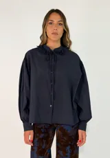 Camisa de algodón color azul marino, con cuello redondo decorado con volados y lazo de ajuste. Presenta mangas largas abullonadas con puños ajustados y cierre frontal con botones.