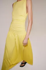 Vestido largo amarillo de algodón, con cuello barco y mangas sisa. Presenta diseño drapeado en los laterales y falda de corte evasé con volumen en el bajo.