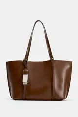 Bolso tipo shopper de color marrón, con diseño minimalista, doble asa de hombro y cierre magnético. Incluye un colgante decorativo con detalle de cinta en tonos claros y un bolsillo interior amplio.