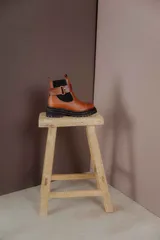 Bota tipo Chelsea de cuero marrón con suela track negra de goma. Presenta paneles elásticos laterales y una costura reforzada en la puntera. Incluye una correa de cuero desmontable con hebilla metálica.