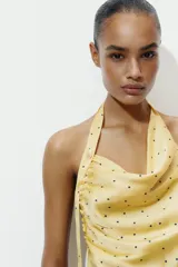 Top de cuello halter con escote fluido y espalda descubierta. Presenta un diseño satinado en color amarillo con estampado de lunares pequeños y un aplique metálico decorativo en el cuello.
