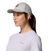 Gorra Columbia Twin Canyon Ball Cap color gris, con visera curva, ojales bordados para ventilación y un parche frontal cuadrado con el logo de la marca en negro, amarillo, marrón y blanco.