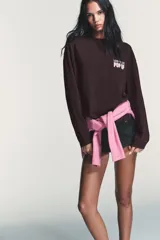 Camiseta de manga larga color marrón oscuro con efecto lavado. Presenta un cuello redondo y un estampado en el pecho con el texto "NEW YORK" en verde y "POP UP" en rosado.