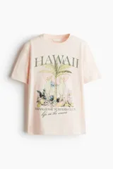 Remera de algodón color rosa pálido, corte holgado, con cuello redondo y estampado frontal de temática hawaiana con palmera, dos loros y texto que dice "HAWAII HANALEI BAY SURFERS CLUB life on the waves".