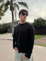 Sweater de punto negro con diseño asimétrico, presentando una mitad lisa y la otra mitad con textura de trenzas. Posee cuello redondo, mangas largas y un calce relajado.