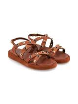 Sandalia marrón con tiras de cuero y tachas doradas y negras.