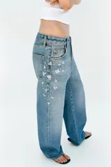 Jeans tiro medio de corte relaxed leg y largo completo, confeccionados en denim azul claro. Presentan detalles de aplicaciones de pedrería y joyas a contraste en los laterales superiores y bolsillos delanteros, estilo clásico de cinco bolsillos y cierre frontal con cremallera y botón metálico.