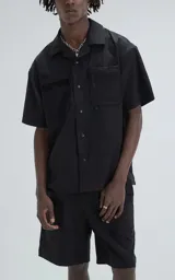 Camisa manga corta de estilo técnico, color negro, confeccionada en microfibra liviana. Presenta cuello camisero, cierre frontal con broches a presión ocultos y doble bolsillo frontal utilitario con detalles de red y cierre.