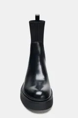 Botín de estilo urbano con suela track, confeccionado en cuero sintético negro con caña de tejido elástico acanalado. Presenta punta redonda, tirador trasero y diseño tipo pull-on.