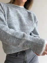 Sweater de punto de corte holgado en color gris melange, con cuello redondo y puños acanalados que presentan un detalle de contraste en color blanco. Posee mangas tipo raglán y una textura suave y elástica.