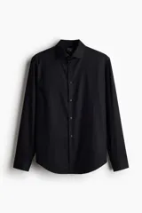 Camisa negra de algodón con cuello italiano, tapeta clásica y canesú en la espalda. Mangas largas con tapeta y botón, y cierre ajustable de botón en los puños. Bajo levemente redondeado. Corte ajustado que realza el contorno del cuerpo creando una silueta entallada.