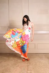 Vestido largo de seda con estampado vibrante y multicolor que incluye motivos frutales y caras sonrientes. Diseño de solera con escote halter anudado al cuello y falda amplia.