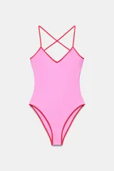 Bañador enterizo de color rosado con detalles de ribetes en color rojo. Presenta escote en pico y tirantes finos cruzados en la espalda.