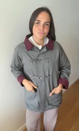 Campera gris de corte recto con cuello y puños en color bordó. Presenta cierre frontal con ganchos metálicos decorativos, bolsillos laterales y forro interno.