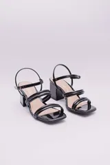 Sandalias de tacón cuadrado medio, confeccionadas en charol negro. Presentan un diseño de tiras finas tubulares en el empeine, punta cuadrada y cierre con pulsera ajustable al tobillo.