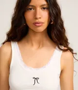 Blusa musculosa confeccionada en tejido rib de algodón con elastano, color blanco, con escote redondo adornado con encaje y detalle de moña bordada en el centro.