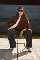 Campera de gamuza color marrón con acabado cepillado suave, cuello clásico, hombros caídos y mangas largas. Cuenta con cierre frontal oculto bajo solapa con botones a presión, bolsillos laterales con cierre y diseño abullonado con elástico interior en el bajo.