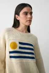 Sweater de lana color crudo con diseño de la bandera de Uruguay tejido en el frente. Presenta un sol amarillo en relieve realizado en crochet y franjas horizontales azules. Posee cuello redondo acanalado y un corte holgado.